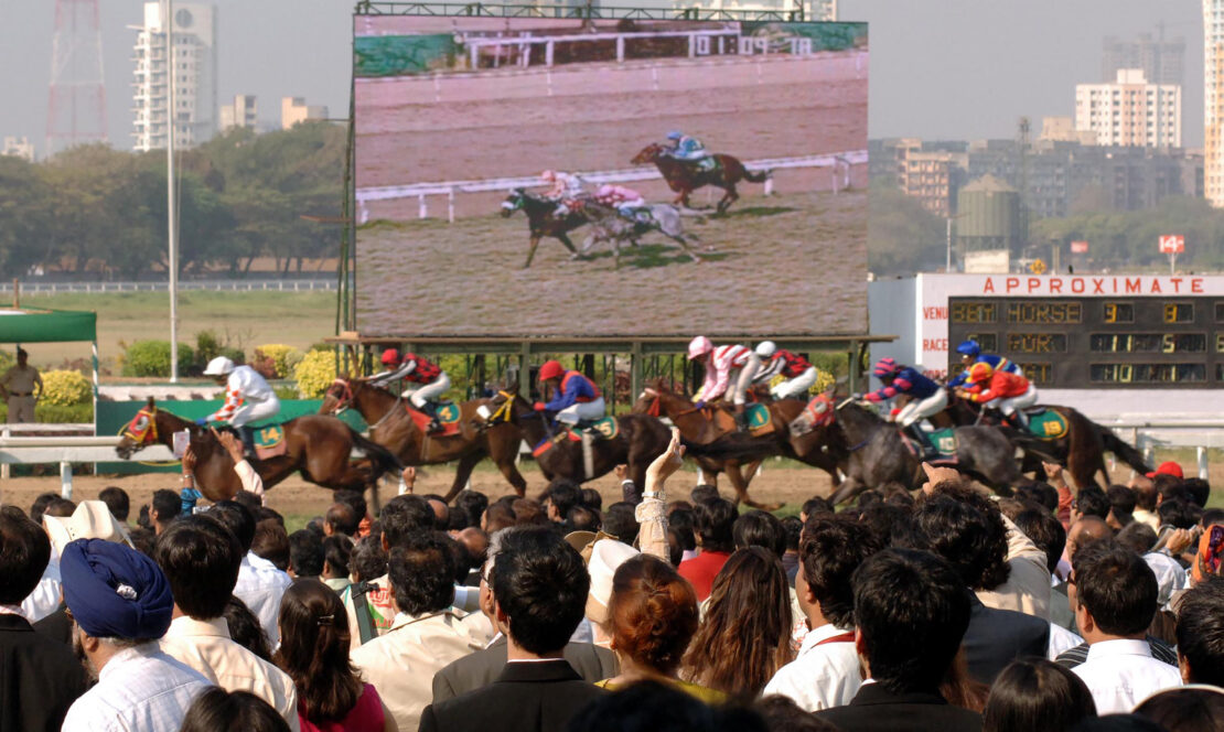 a crowd watching a live race in front of them with a large screen behind the race to help get Indian horse racing explained for both fans and those who might want to bet